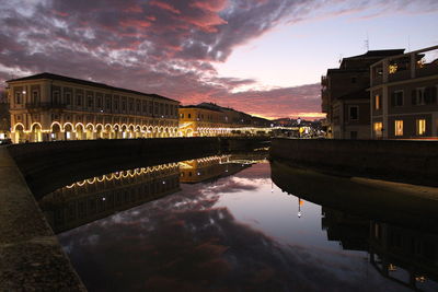 Illuminated buildings by lake against sky at sunset