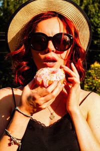 Portrait of woman wearing sunglasses and hat eating donut in park