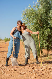 Females farmers in sunhat hugging each other while using rake on ground of field working together in village in summer