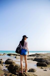 Full length of woman standing on rock at beach against clear sky