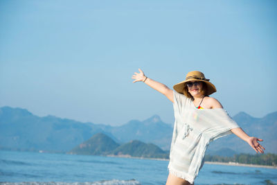 Portrait of young woman with arms outstretched standing at beach by mountains against clear sky