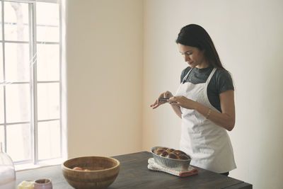 Woman in apron photographing baked challah bread with mobile phone on table against wall at home