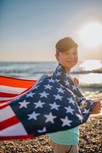 Boy holding american flag at beach against sky