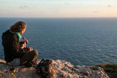 Side view of mature woman playing guitar while sitting on rock against sky during sunset