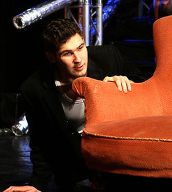 Young man by brown armchair on stage