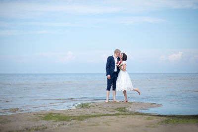 Newlywed couple kissing on beach against sky