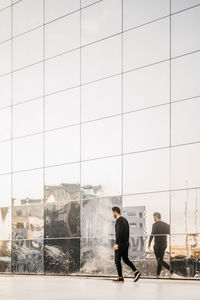 Full length side view of young man walking by reflection on glass building