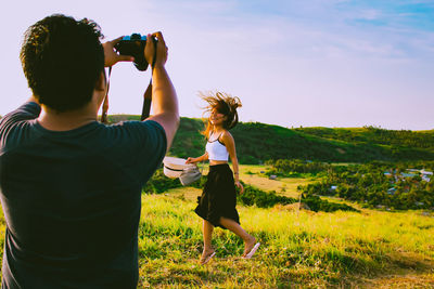 Rear view of young man photographing happy woman walking on field against sky