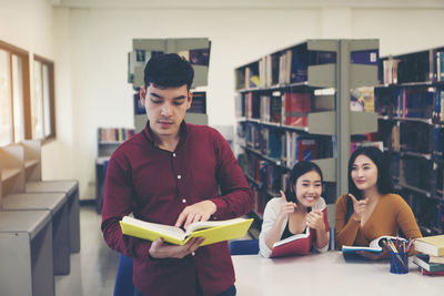 Young man reading book while friends pointing at him in library