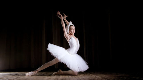 In rays of spotlight, on the stage of the old theater hall. young ballerina in suit of white swan