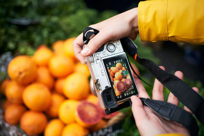 From above of crop anonymous person with digital photo camera taking picture of ripe orange fruits while visiting grocery market