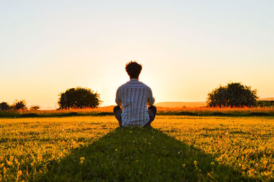 Rear view of man sitting on field against sky during sunset