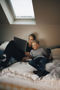 Smiling siblings using laptop while reclining on bed at home