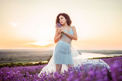 Woman standing on field with purple flowers against sky during sunset