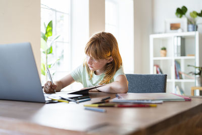 Concentrated redhead girl doing homework in front of laptop at home