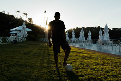 Man with soccer ball on grassy field against sky during sunset