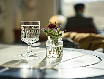 Glass of mineral water, small bouquet of flowers in jar on table in sunlight and silhouette of man