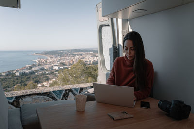 Young woman inside vehicle interior using laptop while sitting on table by sea