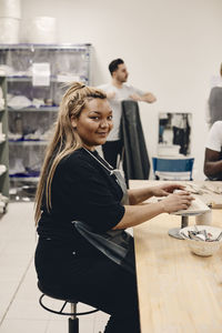 Portrait of smiling young woman with long blond hair molding clay while sitting at table in art class