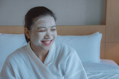 Smiling young woman with facial mask sitting on bed at home