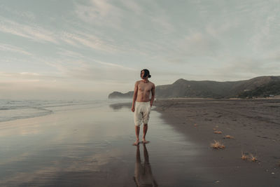 Rear view of man standing at beach against sky during sunset