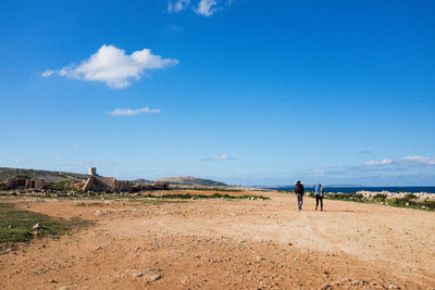 Rear view of friends walking on dirt road against blue sky