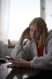 Pensive teenage girl sitting at table and using phone