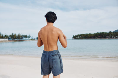 Rear view of shirtless man standing at beach against sky