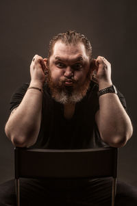 Portrait of man making funny face while sitting on chair against black background