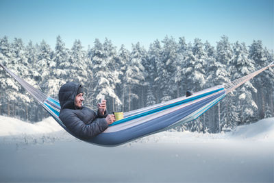 Side view of man using mobile phone while lying down in hammock during winter