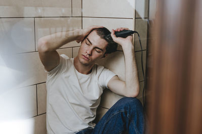 Depressed young man sitting with eyes closed shaving hair