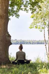 Young woman practicing yoga in nature. sitting on green grass next to a tree and a forest lake. 
