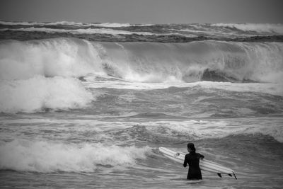 Rear view of woman holding surfboard against waves in sea at beach
