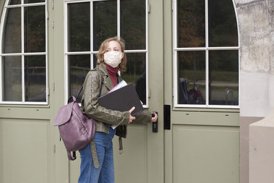 Female student in protective mask holds notes and opens the door to the college