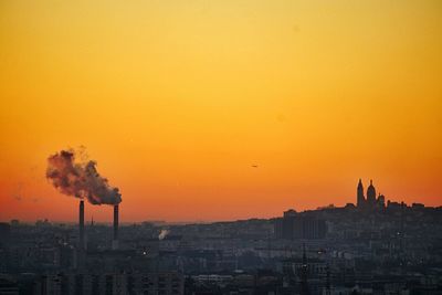 Smoke emitting from industry in city against orange sky during sunset