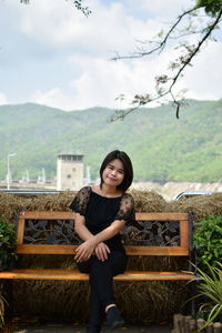 Portrait of smiling young woman sitting on bench against mountains