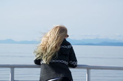 Rear view of teenage girl with blond hair looking at sea while standing by railing against sky