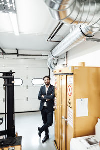 Smiling businessman standing with arms crossed while leaning on orange machinery at industry