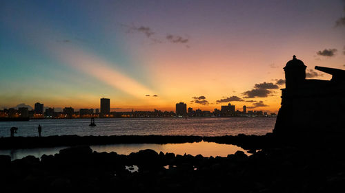 Silhouette buildings by sea against sky during sunset