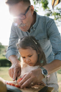 Grandfather teaching granddaughter to cut tomato at table in backyard