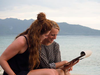 Friends looking at book while sitting by sea against sky