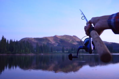 Cropped hand holding fishing rod in lake against sky