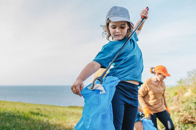 Adorable little volunteer in uniform collecting trash in plastic bag with stick in countryside on summer day