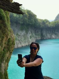 Happy woman taking selfie while standing on cliff against turquoise sea