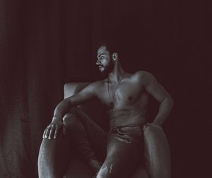 Young man looking away while sitting against black background