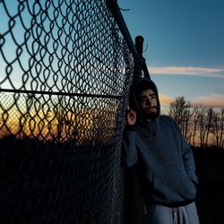 Portrait of man standing by fence against sky during sunset