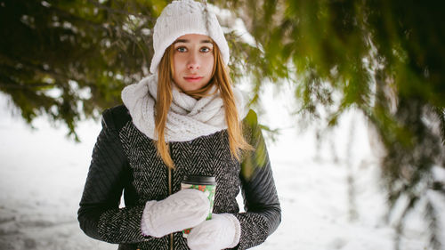 Young woman wearing warm clothing while holding disposable cup amidst trees during winter