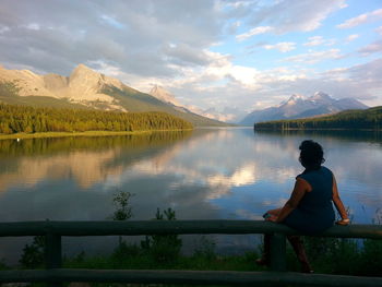 Rear view of woman sitting on railing by lake against mountains