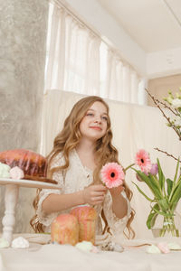 A girl with long hair in a light dress is sitting at the easter table with cakes, spring flowers 