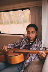 Brown-haired teen girl in plaid shirt playing guitar in camping trailer